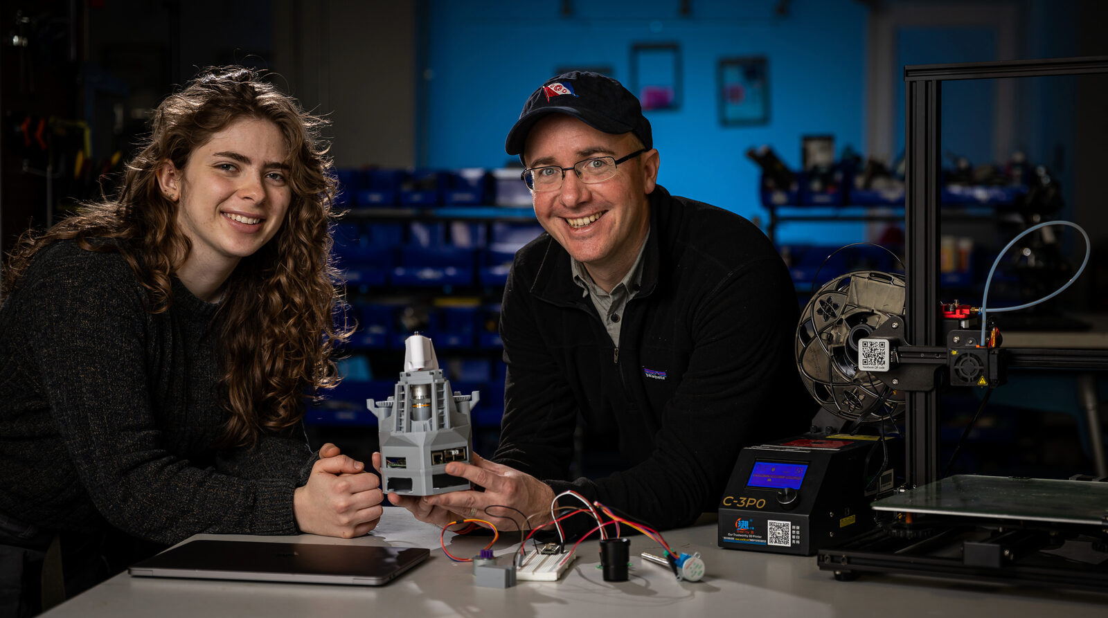 Two people at a workbench with a laptop, a breadboard, and a 3D printer