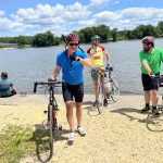 Three riders in helmets pausing with road bikes on a gravel riverbank, steel truss bridge spanning the river behind them